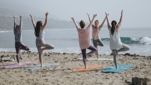 group of women performs standing yoga poses by the beach